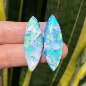 May include: Two oval-shaped opals with a vibrant display of colors. The opals feature a mix of blues, greens, and whites, creating a cloudy, ethereal effect. The stones are polished and smooth, showcasing their natural beauty. The opals are held against a blurred green background.