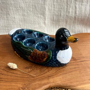 May include: A ceramic duck-shaped egg holder. The duck has a glossy black head, a yellow beak, and a white chest. The body is blue, green, and brown, with space for six eggs. A decorative kitchen item.