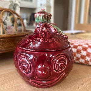 May include: A glossy, deep red ceramic lidded jar shaped like a beetroot with a smiling face. The lid features a green and red stem handle and the word BEETROOT embossed on the top. The jar has a rounded body with sculpted eyes and a mouth.