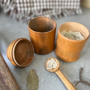 May include: A collection of wooden kitchen items, including three small wooden pots, a wooden spoon holding salt, and a bay leaf. The pots are cylindrical and of varying sizes. The spoon and pots are made of light brown wood.