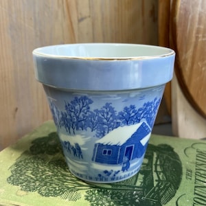 May include: A light blue ceramic planter with a gold rim. The planter features a blue and white winter scene with a cabin and the text "HOME IN THE WILDERNESS". The planter is sitting on a green book.