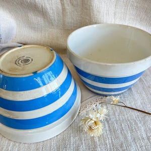 May include: Two white ceramic mixing bowls with blue and white horizontal stripes. One bowl is upright, the other is upside down, revealing the base. The bowls are on a textured, off-white surface.