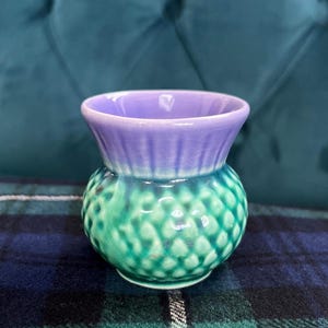 May include: A small ceramic vase with a thistle design. The vase has a textured, green, bulbous base and a smooth, purple rim. The vase is displayed on a plaid surface.