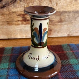 May include: A ceramic candlestick holder with a brown top and base, and a cream-coloured body. The body is decorated with blue, green, and brown floral designs. The words "Guid mich" are written on the base.