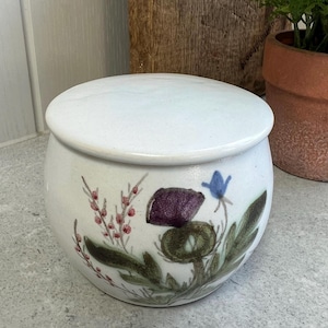 May include: A white ceramic jar with a matching lid. The jar is decorated with a floral design featuring purple thistles, green leaves, and red berries. The jar is round and sits on a gray surface.