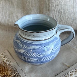 May include: A ceramic jug with a light blue and white wave design. The jug features a spout and a handle. The interior is a darker blue. The jug is placed on a white cloth.