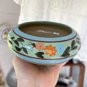 May include: A round ceramic bowl featuring a light blue exterior band with a hand-painted floral pattern. The design includes dark green leaves, small blue dots, and flowers in shades of peach, orange, and light blue, bordered by thin brown lines.