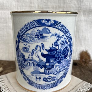 May include: A white ceramic container with a gold rim and a blue and white Asian-inspired design. The design features a pagoda, bridge, and landscape scene. The container is sitting on a white lace doily.