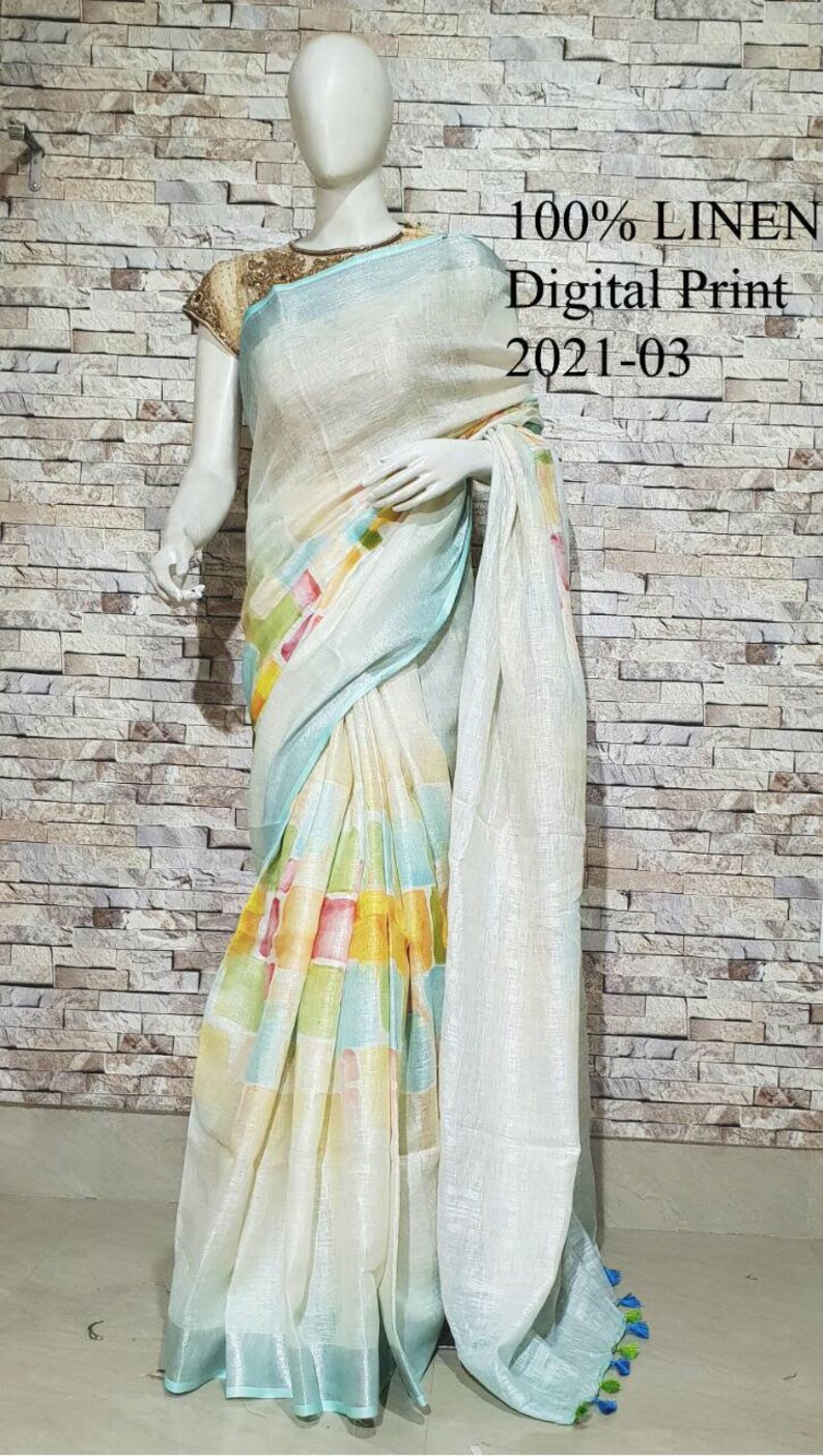 May include: A white linen saree with a blue and multi-colored geometric print. The saree has a silver shimmer and a blue border. The text "100% LINEN Digital Print 2021-03" is visible in the image.
