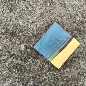 Puede incluir: Un posavasos cuadrado de madera con una parte superior de resina azul y una base de madera natural. La resina tiene un efecto veteado con tonos azules y blancos, y una fina franja marrón separa la resina de la madera.