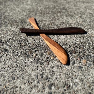 May include: Two wooden butter knives, one is light brown and the other is dark brown. The knives are crossed on a gray concrete surface.