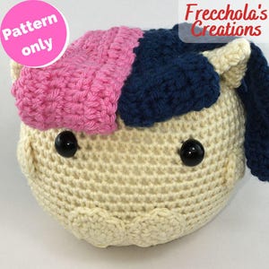 Può includere: Una testa di animale all'uncinetto in crema, rosa e blu navy. La testa ha occhi neri a bottone e un cerchio rosa con le parole "Pattern only". Il testo "Frecchola's Creations" si trova nell'angolo in alto a destra.
