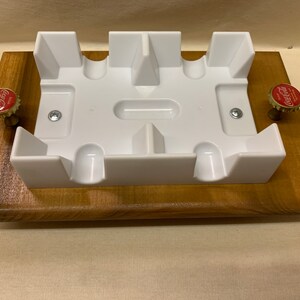 May include: A white plastic card holder with slots for holding cards. The holder is mounted on a wooden base with two bottle caps on the sides.