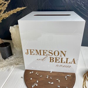 Op de afbeelding: Witte acryl bruiloft kaartenbox met een gleuf aan de bovenkant. De doos heeft gouden letters met de tekst "Jemeson en Bella 10.11.2022".