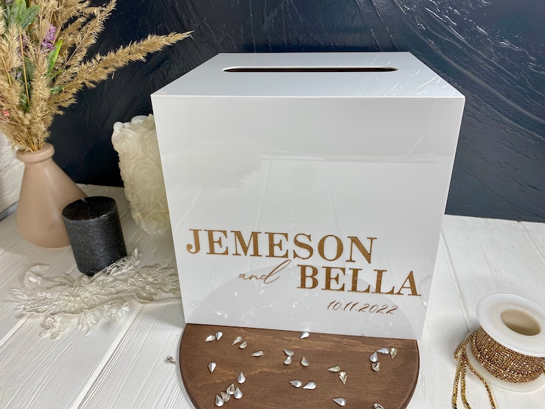 Op de afbeelding: Witte acryl bruiloft kaartenbox met een gleuf aan de bovenkant. De doos heeft gouden letters met de tekst "Jemeson en Bella 10.11.2022".