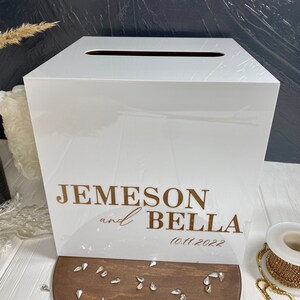 Op de afbeelding: Witte acryl bruiloft kaartenbox met een gleuf aan de bovenkant. De doos heeft een gouden inscriptie met de tekst "Jemeson en Bella 10.11.2022".