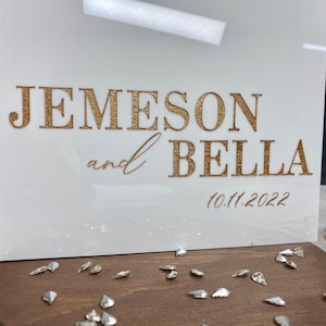 Op de afbeelding: Een wit acryl bord met gouden letters waarop "Jemeson en Bella" staat, met daaronder de datum "10.11.2022".