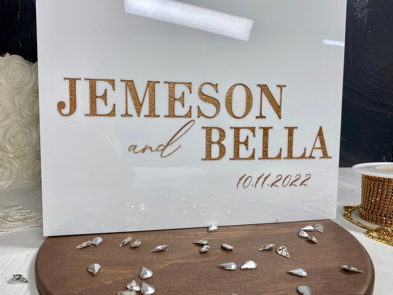Op de afbeelding: Een wit acryl bord met gouden letters waarop "Jemeson en Bella" staat, met daaronder de datum "10.11.2022".