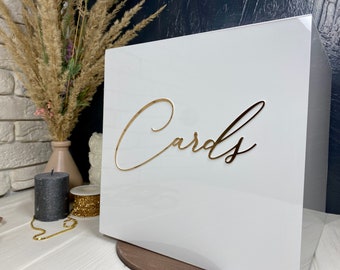 Könnte beinhalten: Weiße Acrylbox mit einer goldenen Aufschrift "Karten".