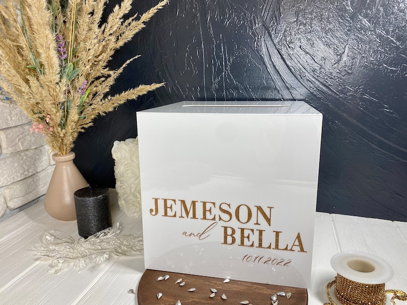 Op de afbeelding: Witte vierkante doos met een gleuf aan de bovenkant. De doos heeft gouden letters met de tekst "Jemeson en Bella 10.11.2022".