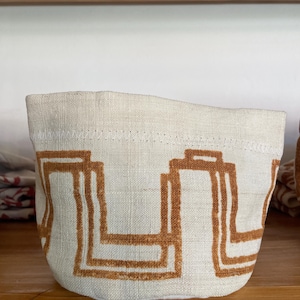 Può includere: Un cestino in tessuto beige con un motivo geometrico marrone. Il cestino ha una forma arrotondata e un bordo superiore cucito. Il design è una serie ripetuta di forme rettangolari. Il cestino è su una mensola di legno.