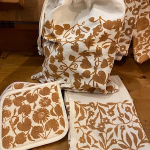 Può includere: Un set di tessuti da cucina in colore naturale con un motivo floreale marrone. Il set include un sacco con coulisse, presine e un canovaccio. Il motivo floreale presenta fiori e foglie stilizzate, creando un set da cucina decorativo.