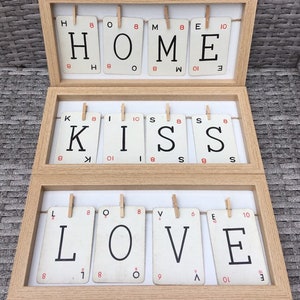 May include: A wooden frame with three rows of Scrabble tiles spelling out "HOME", "KISS", and "LOVE". The tiles are held in place with clothes pegs.