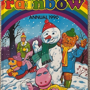 May include: The cover of a "Rainbow Annual 1990" book, showing a rainbow and cartoon characters like a snowman and a bear in a snowy scene. The title "rainbow" is in bright, multi-colored letters.