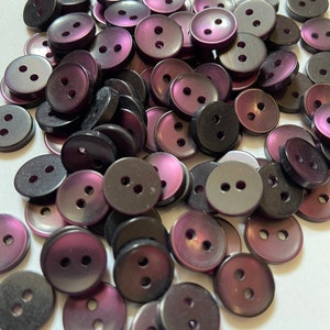 May include: A collection of two-hole buttons in shades of black and purple. The buttons have a glossy finish and are arranged in a random pattern.