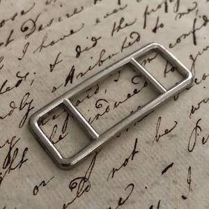 May include: A silver metal buckle, rectangular in shape with a central bar. The buckle is displayed on a backdrop of aged paper with handwritten script. The design of the buckle is simple and geometric.