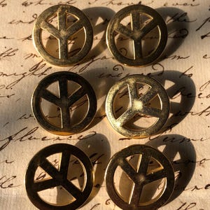 May include: Six gold-coloured peace sign buttons on a vintage paper background. The buttons are circular with a classic peace symbol design. The paper has handwritten text.
