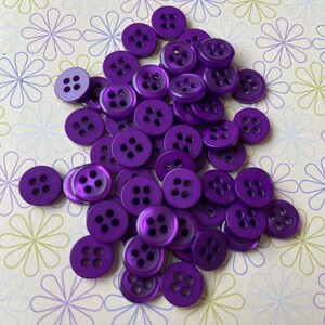 May include: A pile of purple plastic buttons with four holes each. The buttons are shiny and have a slight sheen.