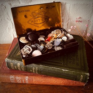 May include: A vintage rectangular metal tin labelled KAY-GEE MEDIUM NAVY CUT CIGARETTES filled with a variety of assorted buttons in different shapes, sizes, and colours including white, black, brown, blue, and metallic tones, resting on two books.