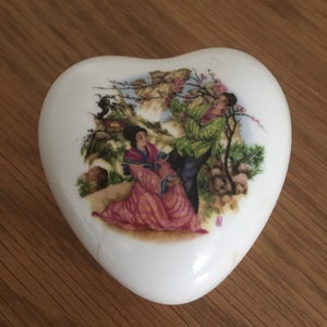 May include: White heart-shaped ceramic trinket box with a painted scene of two people in traditional clothing standing in a garden with a tree and flowers.