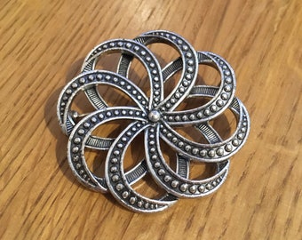 May include: A silver-toned metal brooch in the shape of a flower with a circular design. The brooch has a detailed, intricate design with multiple layers of circles and dots.
