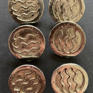 May include: Six silver-colored metal buttons with a wavy design. The buttons are round and have a raised edge.