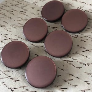 May include: Six circular, mauve-coloured buttons with a glossy finish are arranged on a vintage paper background. The buttons are evenly spaced, showcasing their smooth, circular design. These buttons are ideal for sewing and craft projects.