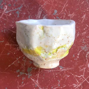 May include: A small white ceramic bowl with a textured, uneven surface. The bowl has a yellow and orange glaze with a speckled pattern.