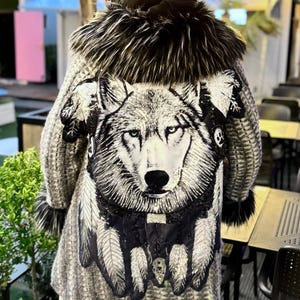 May include: A grey and white patterned coat with a large wolf graphic on the back. The coat has a black fur collar and trim, and is worn with a black hat. The wolf design includes feathers and beadwork.