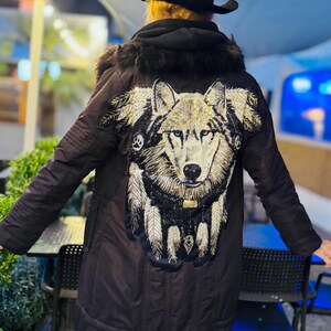 May include: A dark brown winter coat with a large embroidered wolf design on the back. The wolf is depicted with intricate detail, featuring gold and silver accents. The coat has a fur-lined hood and a black hat.