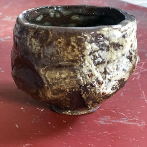 May include: Handmade ceramic bowl with a rough, textured surface. The bowl features a dark brown interior and exterior with patches of beige and white. The rim is uneven, adding to the rustic aesthetic. The bowl sits on a red surface.