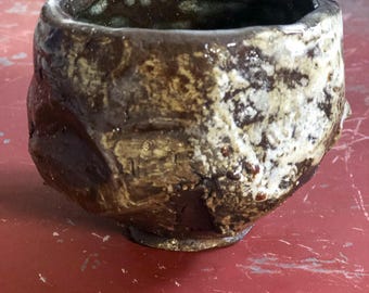 May include: Handmade ceramic bowl with a rough, textured surface. The bowl features a dark brown interior and exterior with patches of beige and white. The rim is uneven, adding to the rustic aesthetic. The bowl sits on a red surface.