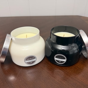 May include: Two Capri Blue Volcano scented candles. One is white, the other is black. Each candle has a silver lid and a label that reads "Capri Blue Volcano". The candles are on a brown wooden surface.