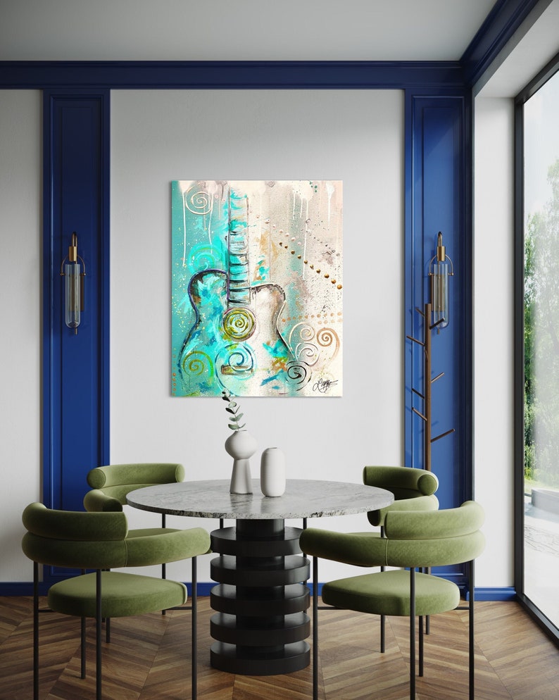 May include: A teal blue acoustic guitar with white and gold accents is painted on a canvas with a white background. The guitar is surrounded by swirls and dots in shades of blue, white, and gold. The painting is hanging on a white wall in a room with a green velvet dining chair and a round marble table.