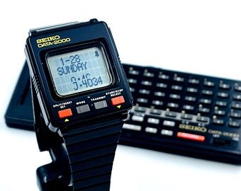May include: A black Seiko Data 2000 digital watch with a black band. The watch face displays the date, day of the week, and time. The watch is on a stand next to a black Seiko Data 2000 calculator.
