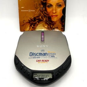 Puede incluir: Un reproductor de CD Sony Discman ESP plateado con borde negro, con una pantalla digital que muestra la hora. La carátula del CD en el fondo muestra el álbum "Frozen" de Madonna.