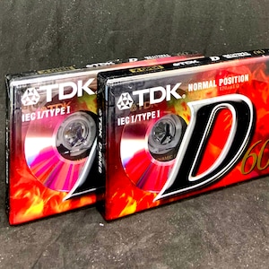 Puede incluir: Dos paquetes sellados de cintas de cassette de audio TDK D-60. Los paquetes son rojos y negros con una cinta de cassette plateada y negra en el interior. El texto en los paquetes dice "TDK" e "IEC I/TYPE I". El texto en el paquete de la derecha también dice "NORMAL POSITION 120MIN/60".