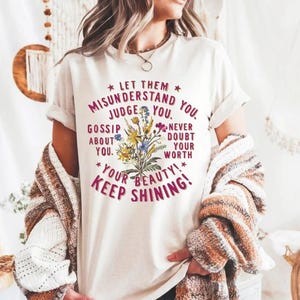 以下が含まれることがあります： 白いTシャツにピンクの花柄のデザインと「誤解させてください。判断してください。あなたについて噂してください。あなたの価値を決して疑わないでください。あなたの美しさ！輝き続けましょう！」というテキストが書かれています。