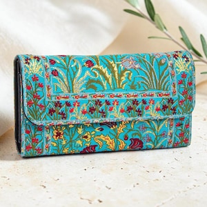 May include: A rectangular wallet featuring a vibrant turquoise background with an intricate woven pattern of red, gold, and green floral and botanical motifs. The design includes a flap closure with a repeating border and detailed plant imagery.