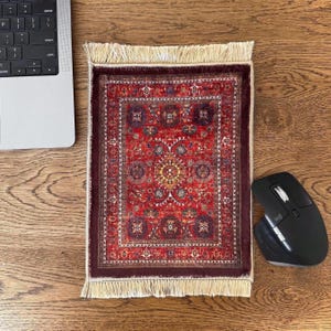 May include: A rectangular mouse mat featuring a detailed red, burgundy, and navy blue oriental rug pattern with a central medallion and intricate floral motifs. The mouse mat has cream-coloured fringe trim along the top and bottom edges.